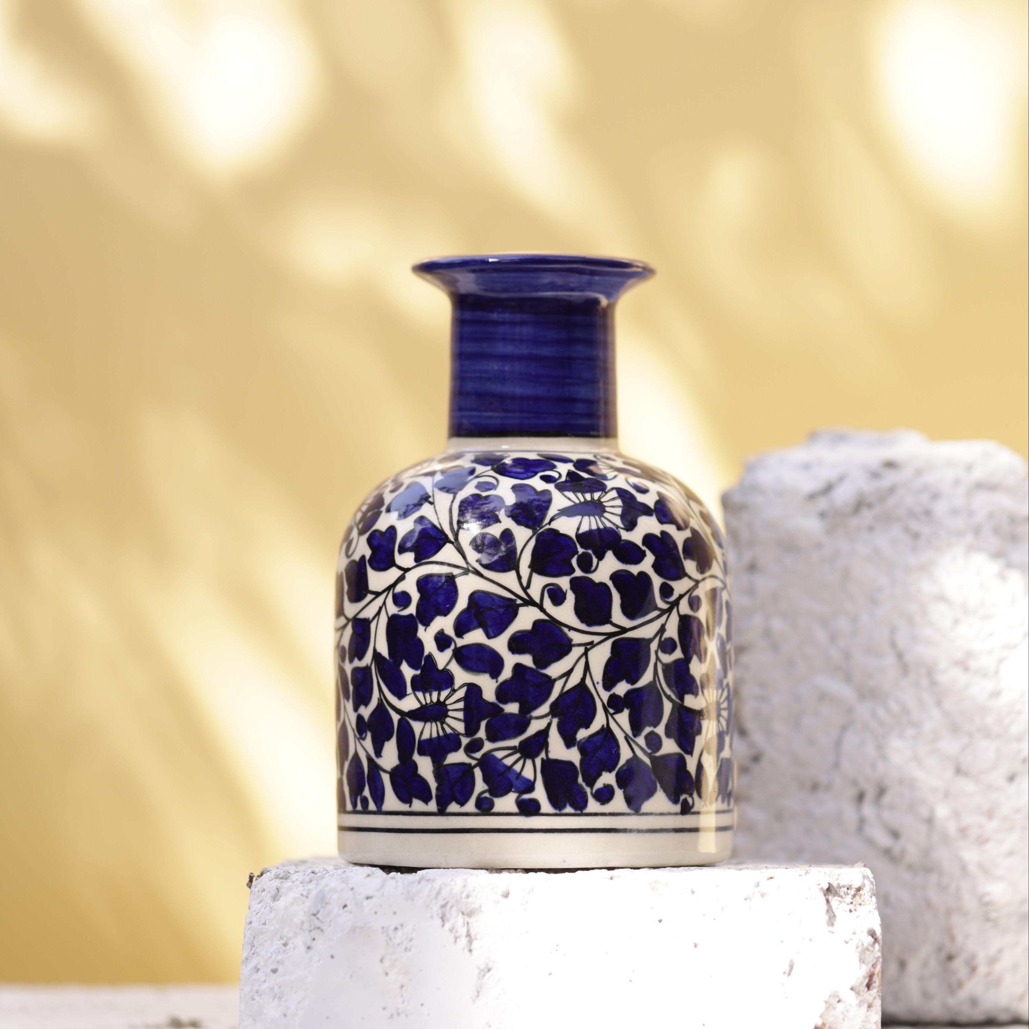 Hand-painted Sphinx ceramic vase, This vase has a distinctive dark blue ridged neck and a body adorned with a detailed floral and foliage design in dark blue and black on a white base. It is displayed on textured white stones and brick.