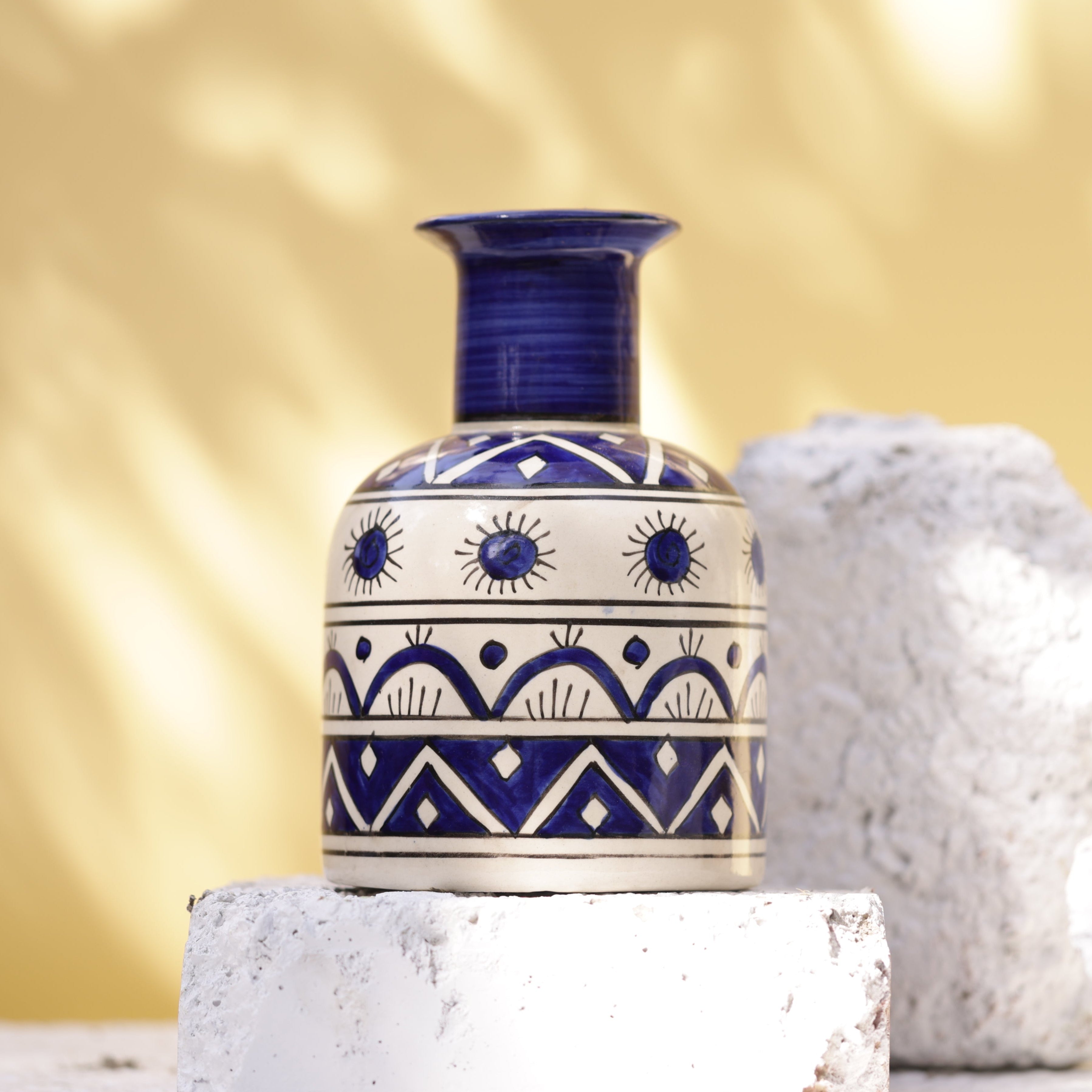 A handcrafted Sphinx ceramic vase in a Moroccan-inspired style. The vase features a dark blue, vertically ridged neck and a rounded body decorated with geometric patterns in dark blue and white, including zig-zag lines, stylized sunbursts, and horizontal bands. The vase rests on a white brick, flanked by two rough, white stones, against a blurred yellow and white background.