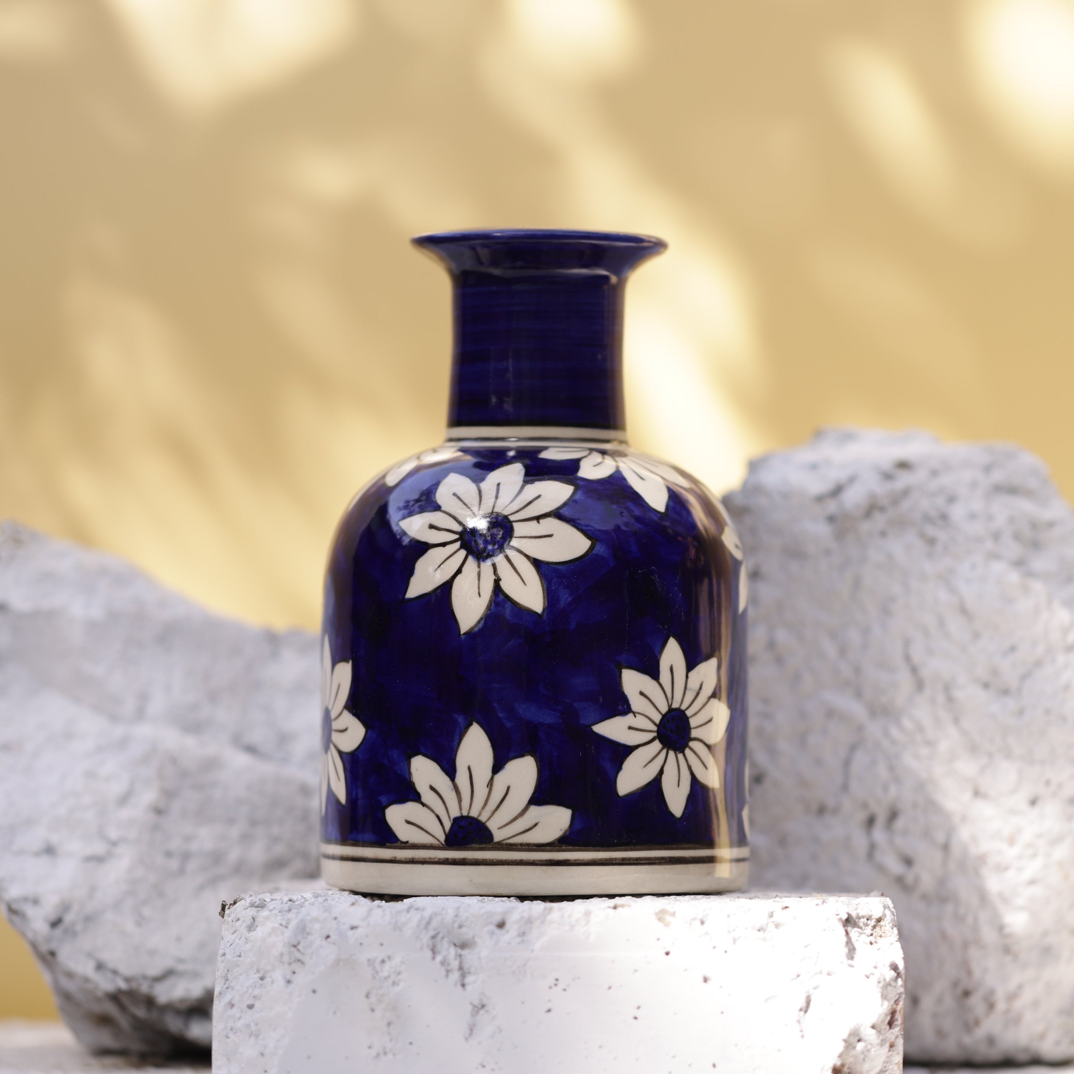 A handcrafted Sapphire Petal Vase. The vase features a deep sapphire blue, vertically ridged neck and a rounded body decorated with large, stylized white daisy-like flowers with dark centers and radiating petals. A thin white band borders the bottom of the patterned area. The vase rests on a white brick, flanked by two rough, white stones, against a blurred yellow and white background.