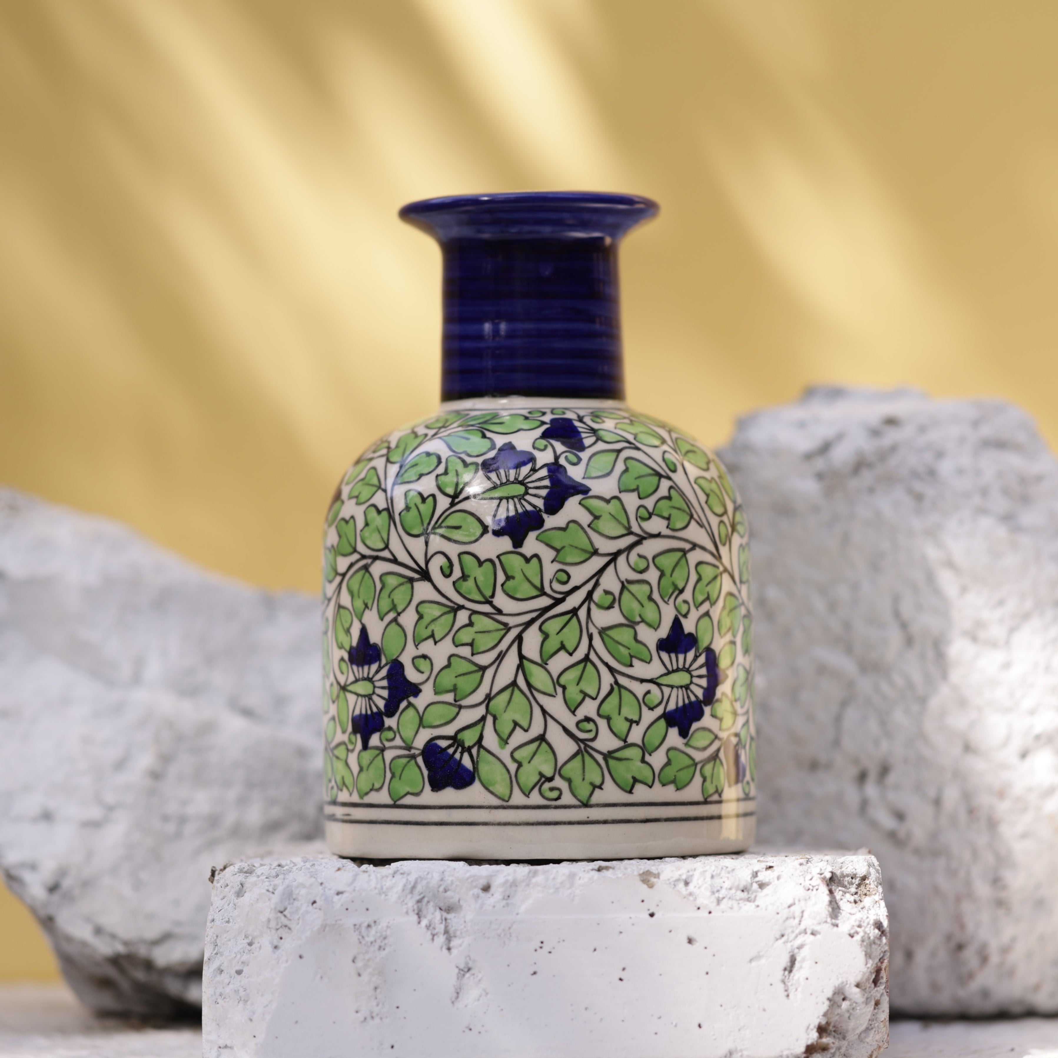 Hand-painted Sphinx ceramic vase, This vase has a distinctive dark blue ridged neck and a body adorned with a detailed floral and foliage design in green and dark blue on a white base. It is displayed on textured white stones and brick.