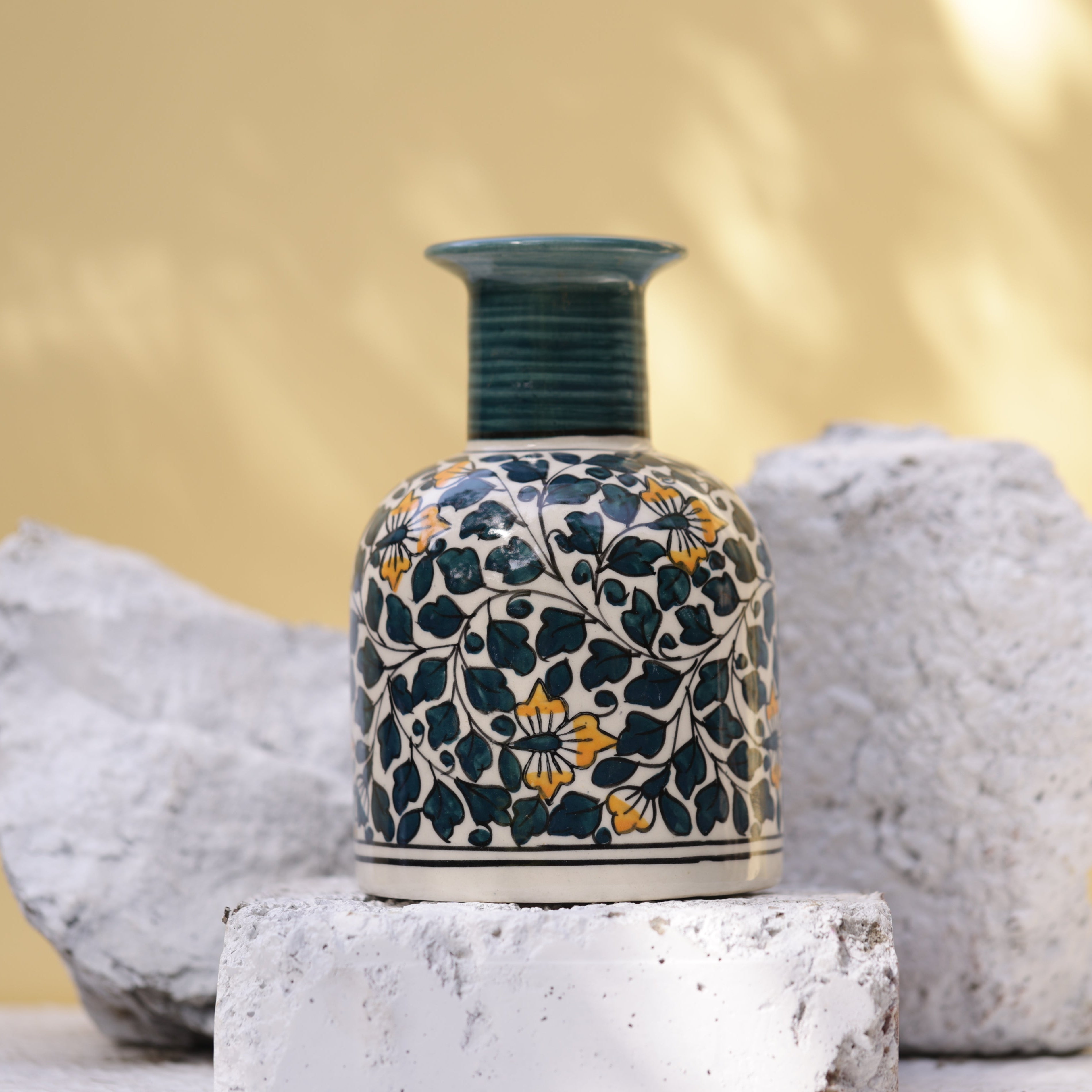 A vibrant ceramic vase, handcrafted is artfully presented on white bricks and stones against a blurred outdoor backdrop. The vase showcases a rich pattern of dark teal leaves and yellow flowers with orange centers on a dark blue background, complemented by a dark teal ridged neck.