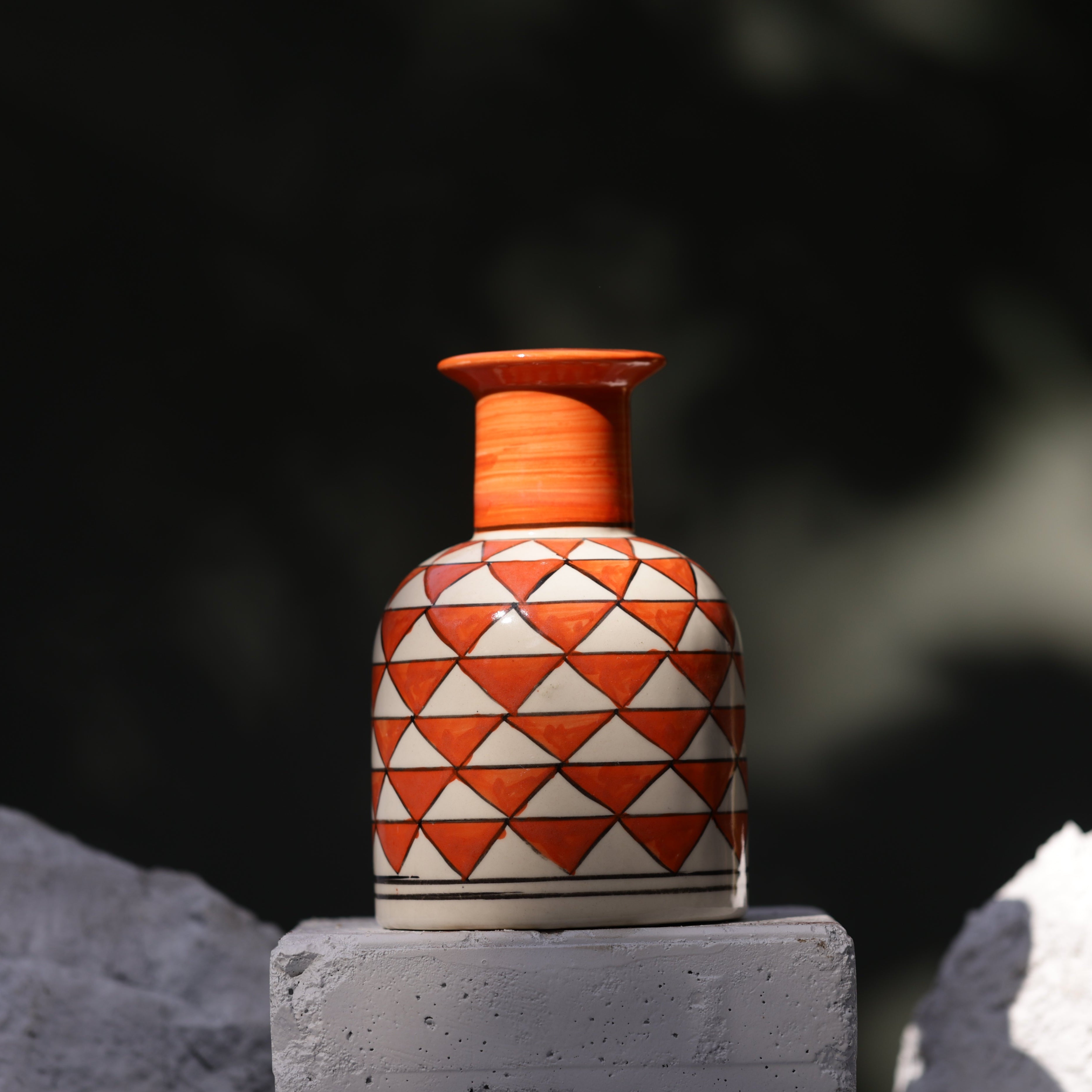 A handcrafted ceramic vase with an orange, horizontally ridged neck and a rounded white body. The body features a pattern of alternating white and orange triangles outlined in black. The vase rests on a stack of white bricks, flanked by two rough, white stones, against a blurred dark background with dappled sunlight.