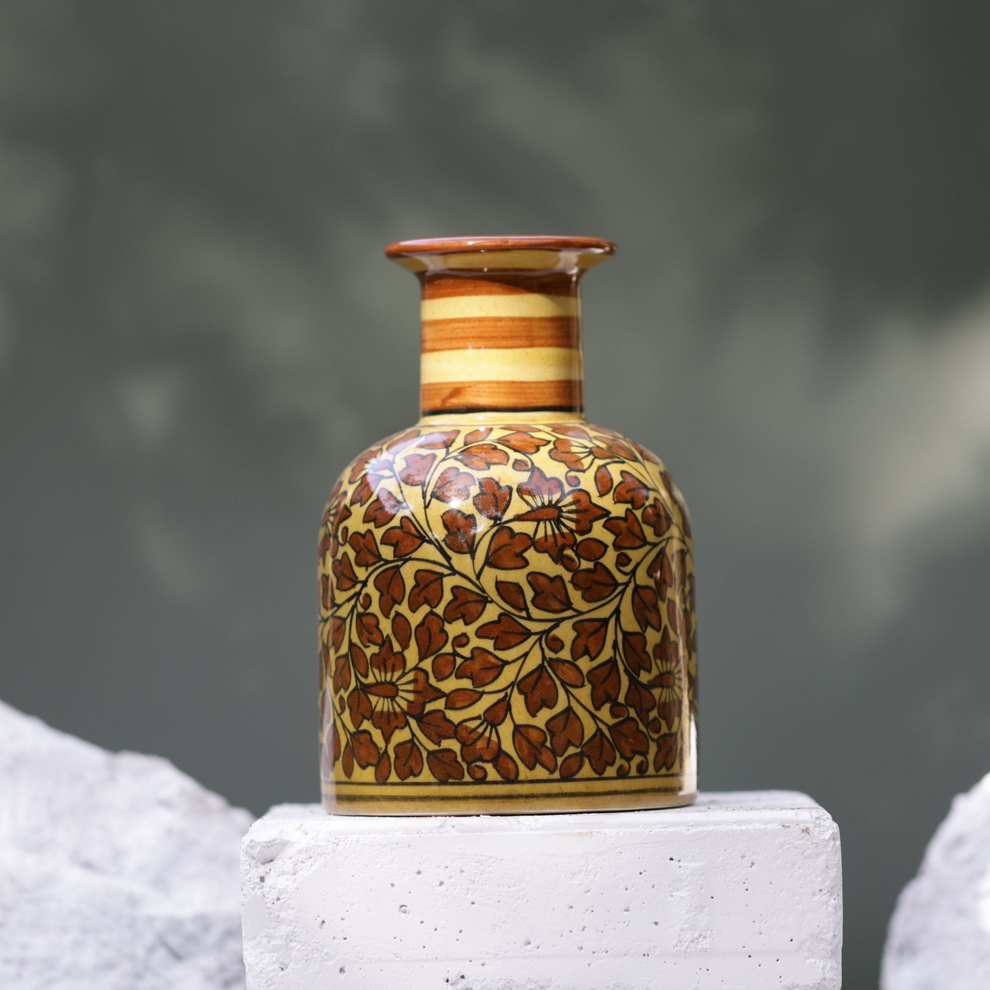 A handcrafted ceramic vase featuring a warm yellow base with intricate brown floral and leaf patterns. The vase has a rounded body and a cylindrical neck with horizontal brown and beige stripes. It rests on a white brick, with two rough, white stones flanking it, against a blurred green and gray background.