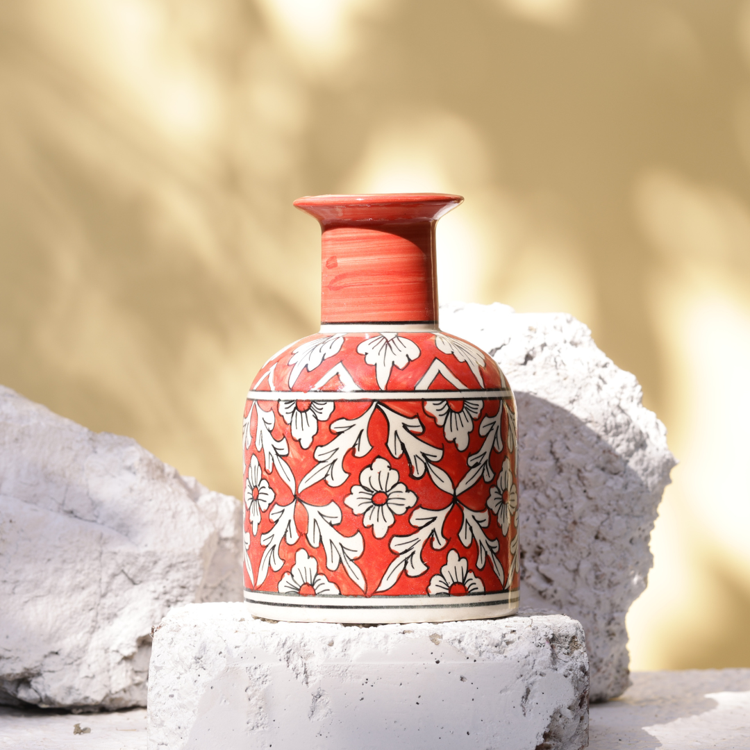 A handcrafted red and white ceramic flower pot . The pot has a rounded body with a narrow neck and a flared rim. It is decorated with hand-painted white floral and leaf patterns on a red background, with black and white striped accents. The pot rests on a rough, white stone or brick, with another similar stone behind it, against a blurred yellow and white background.